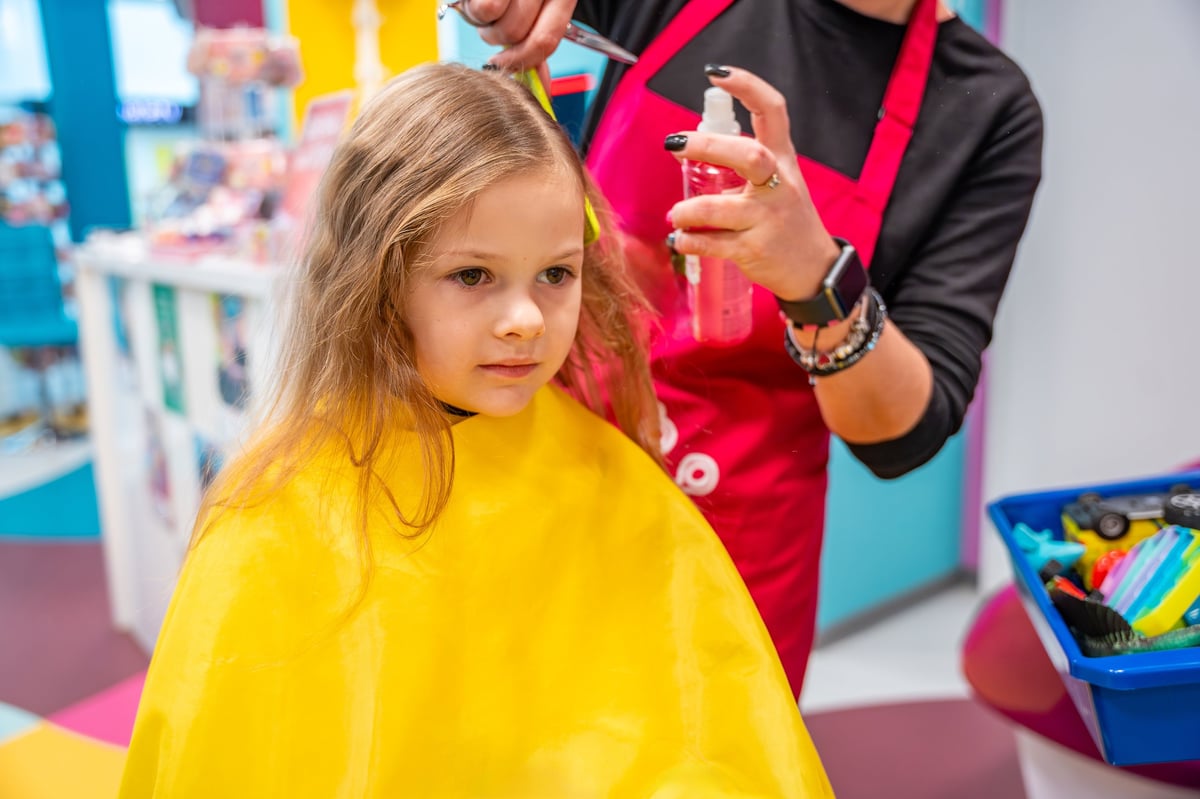 Hairdresser making a hair style to cute little girl in real child salon with colorful interior and toys