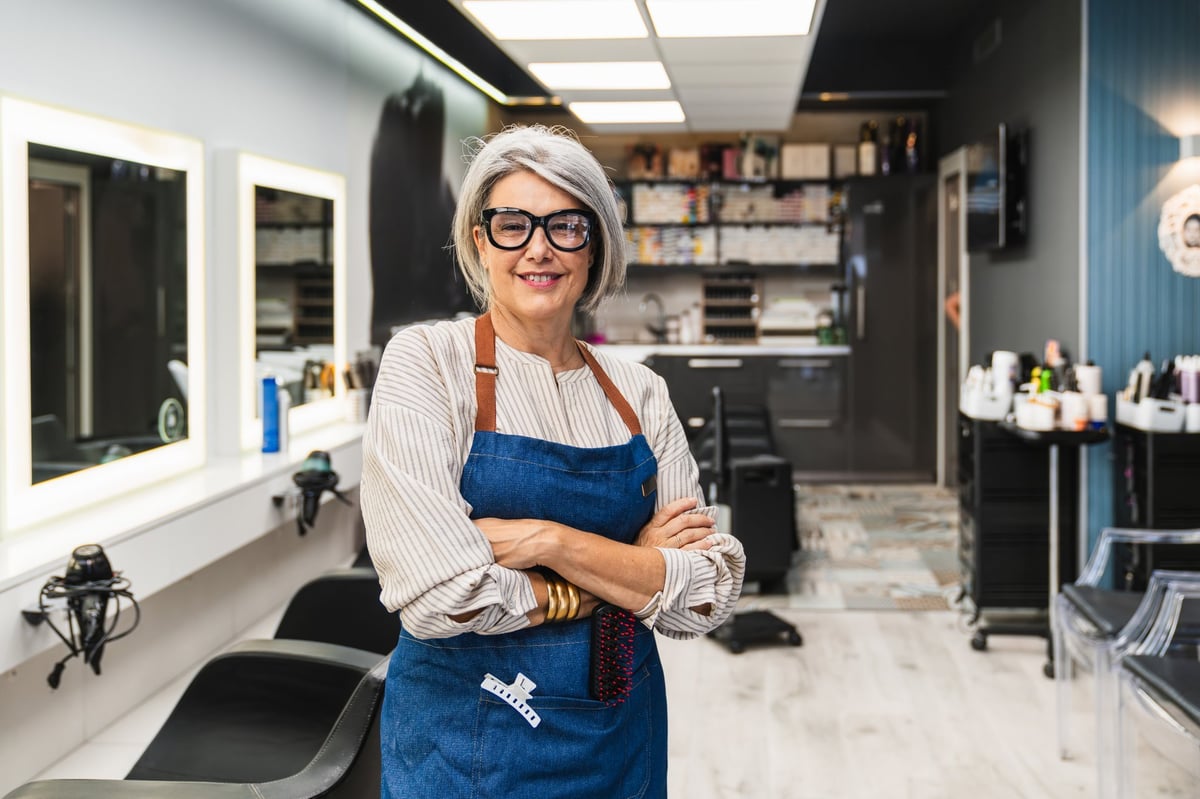 Confident businesswoman with gray hair and glasses standing with arms crossed in her modern hair salon
