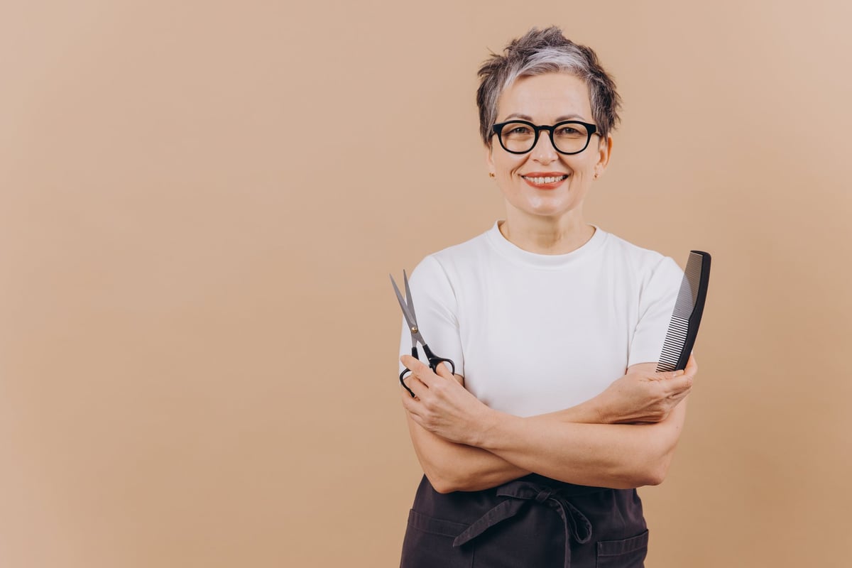 Smiling woman hairdresser holding scissors and comb, wearing an apron and glasses, ready to provide hairstyling and cutting services