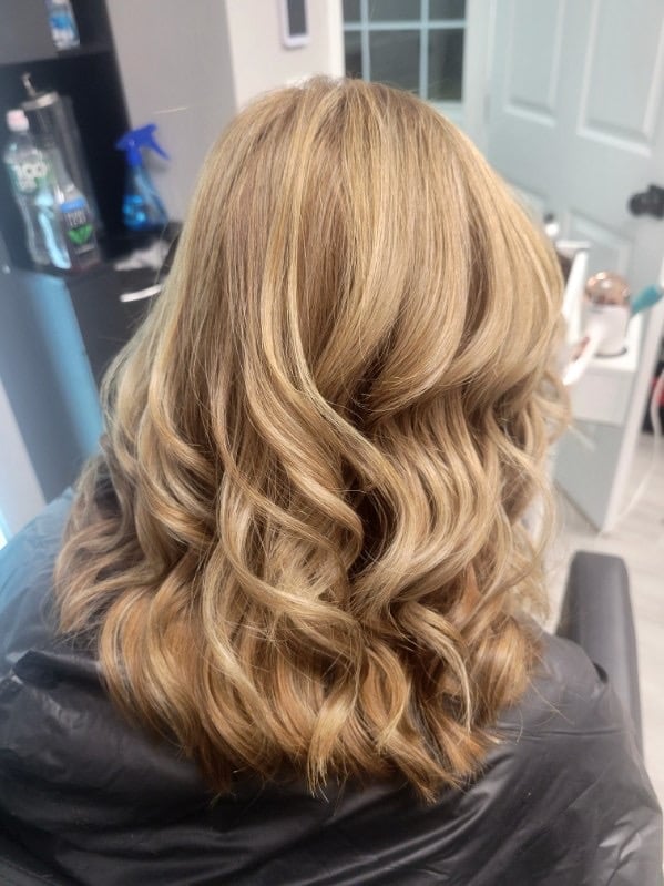 Back view of a woman with shoulder-length, wavy golden blonde hair styled with loose curls in a salon setting