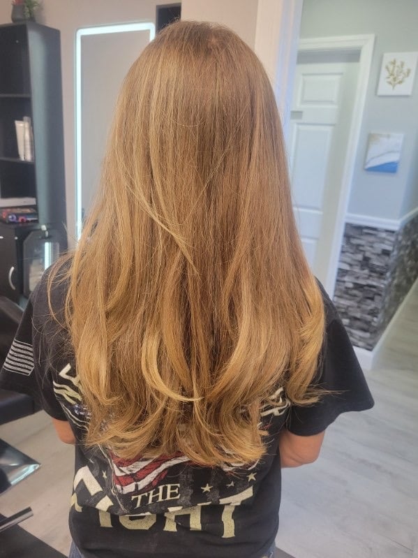 Back view of a person with long, layered honey-blonde hair wearing a black graphic t-shirt in a salon setting