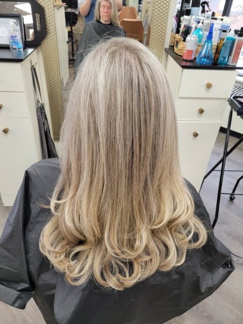 Woman with long blonde wavy hair in a salon chair, back view showing layered haircut and styling