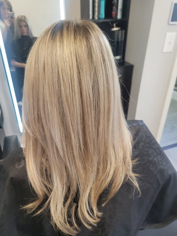 Back view of a woman with long, layered blonde hair styled straight, in a salon mirror setting